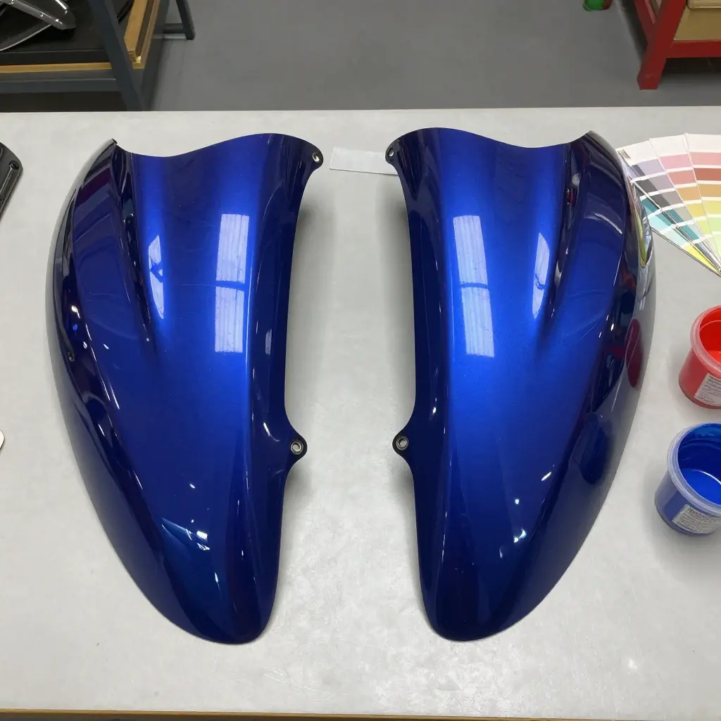 Two 600cc sportbike fairings side by side on a workbench, showing a factory metallic pearl blue panel next to an aftermarket panel in a slightly different shade, with a paint swatch fan nearby